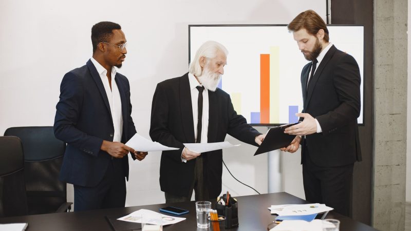 Three businessmen discussing documents in a conference setting with a presentation screen. — Littlepepe Presale: Revolutioniz