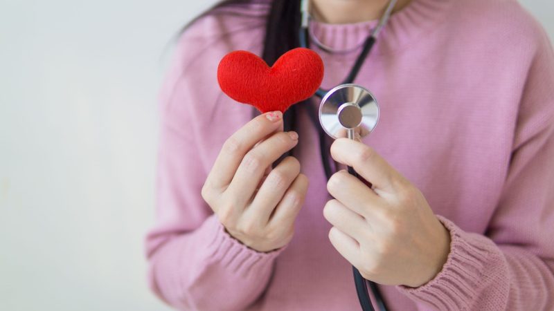 Unrecognizable female in pink sweater with stethoscope on neck standing on white background with red heart in hand in dayligh